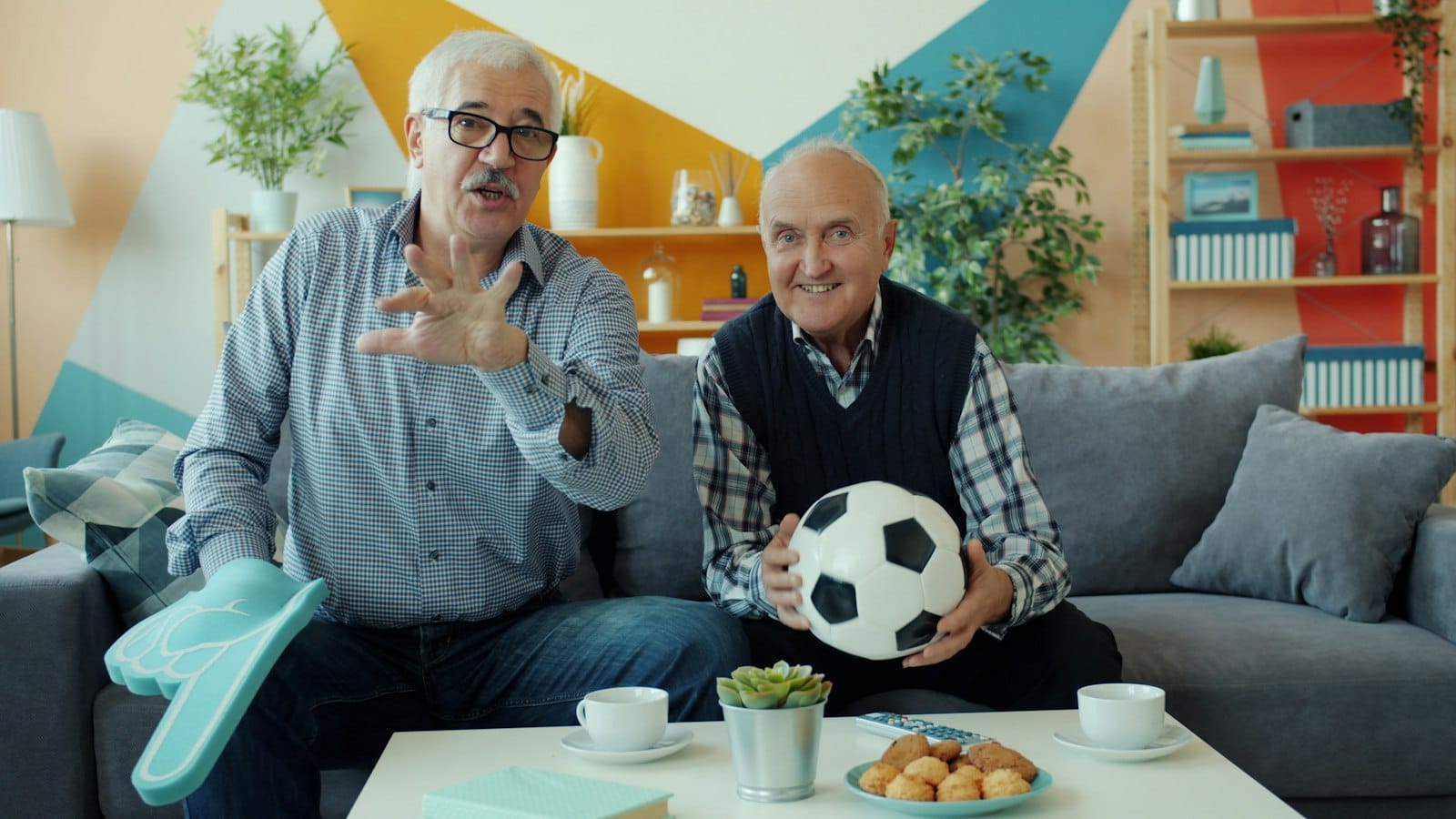 Two friends on a sofa cheering a match, one holding a soccer ball